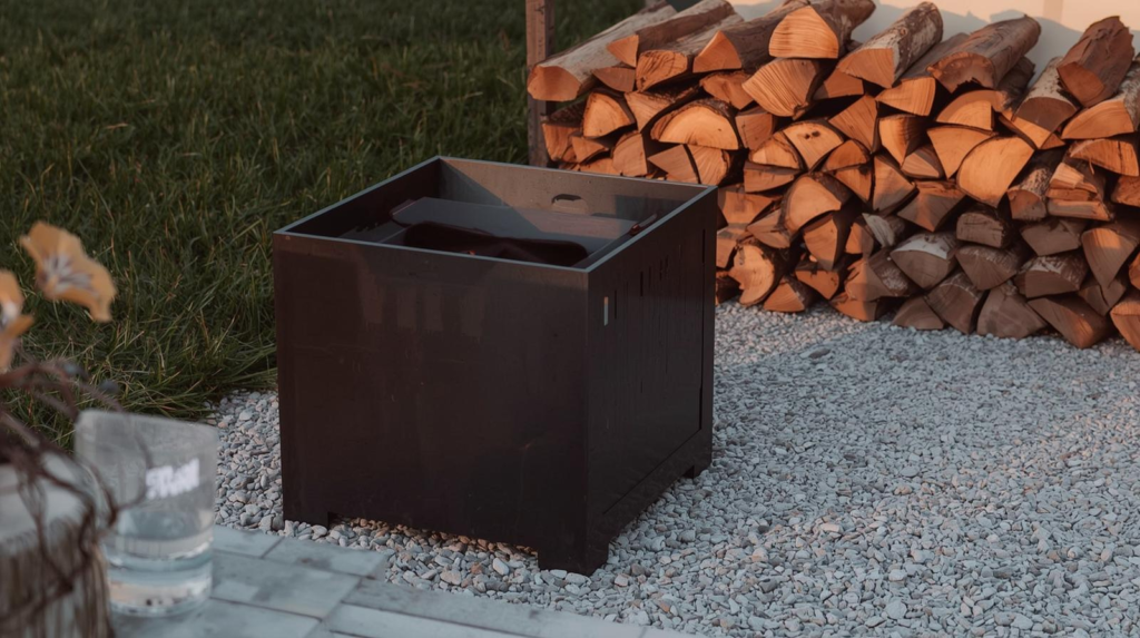 Modern steel fire pit on a gravel patio with a small stack of firewood in warm evening light.